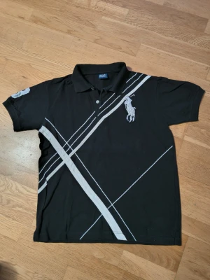 Svart pikétröja från Ralph Lauren - Svart pikétröja från Ralph Lauren med vita diagonala ränder och stor broderad logga på bröstet. Kragen har tryckt text och ärmarna har siffran 3 i vitt. Klassisk pikékrage och korta ärmar, perfekt för en sportig look.