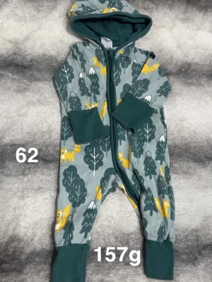 Grön jumpsuit med skogsmönster - Mysig jumpsuit i grönt med huva och dragkedja framtill. Mönstrad med mörkgröna träd och gula rävar, samt breda muddar vid ärmar och ben. Perfekt för chill dagar hemma eller när du vill vara extra bekväm.🌸 Fint använt skick. Storlek 62