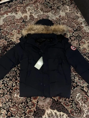 Svart Canada Goose dunjacka med päls - Säljer en svart dunjacka från Canada Goose med ikonisk patch på ärmen och stor huva med fluffig päls. Jackan har flera fickor, dragkedja och knappar framtill. Perfekt för kalla dagar och riktigt snygg till vinterlooken.