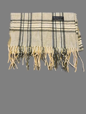 Rutig beige scarf från Burberrys - Säljer en klassisk rutig scarf från Burberrys i beige och grå toner, tillverkad i 100% lammull. Scarfen har fransar längs nederkanten och ett stilrent mönster som ger en tidlös vibe. Perfekt för dig som gillar brittisk design och vill ha något extra i garderoben. Perfekt nu eftersom snön har kommit❄️Kom med frågor eller prisförslag!