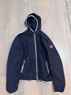 Mörkblå vindjacka med huva Colmar - Snygg mörkblå vindjacka från Colmar med vit dragkedja och detaljer. Jackan har huva, två fickor med dragkedja och vit kant längs huvan och nederkanten. Perfekt för blåsiga dagar och enkel att slänga på sig.