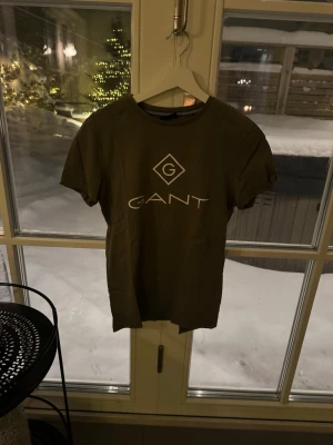 Olivgrön GANT t-shirt XS - Snygg olivgrön t-shirt från GANT i storlek XS. Klassisk passform med korta ärmar och rund hals. Stor GANT-logga i vitt framtill. Tillverkad i mjuk bomull som känns skön mot huden.