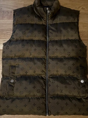 Louis Vuitton dunväst med monogram -  KAN GÅ NER I SNABBT KÖP Säljer en mörkbrun dunväst från Louis Vuitton med ikoniskt monogrammönster över hela västen. Västen har ståkrage, två fickor med tryckknappar och dragkedja framtill. Materialet är bomull och polyester, vilket gör den både mjuk och slitstark.