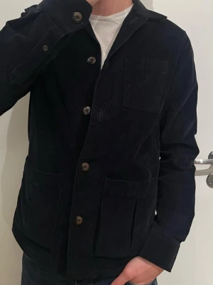 Svart manchester overshirt från John Henric - Mörkblå overshirt från John Henric i 100% manchester med klassisk krage, stora frontfickor och knappar framtill. Perfekt att slänga på över en t-shirt för en chill och stilren look. Snygg struktur och skön passform.