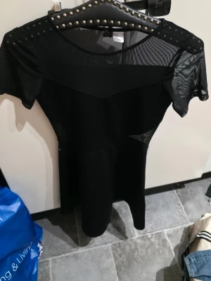 Svart meshklänning från Divided - Svart kortklänning från Divided med meshdetaljer upptill och på ärmarna. Klänningen har rund hals och är kortärmad, med en tight passform. Materialet är 95% polyester och 5% elastan vilket gör den stretchig och bekväm. Perfekt för dig som gillar en stilren look.