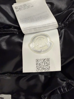 Svart Moncler pufferjacka strl 2 - Säljer en svart pufferjacka från Moncler i storlek 2. Jackan har en glansig finish och är tillverkad i nylon med dunfyllning. Märket syns tydligt på innerlappen och medföljande tags och box ingår. Perfekt för dig som vill ha en lyxig och varm jacka.