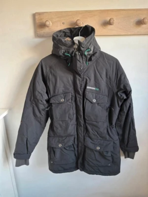 Svart parkajacka från Didriksons - Svart parkajacka från Didriksons med huva och gröna detaljer på snörningen. Jackan har flera stora fickor med knappar, dragkedja framtill och logga på bröstet. Perfekt för kalla dagar och har en schysst streetstil.