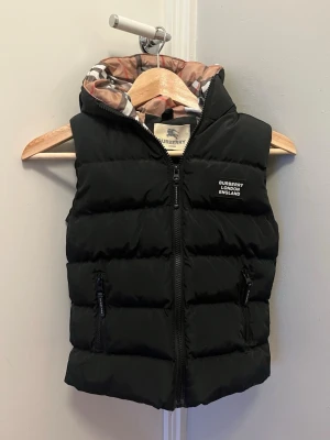 Svart dunväst från Burberry för barn - Säljer en svart dunväst från Burberry med klassiskt rutigt foder i beige, vitt och rött. Västen har huva, två fickor med dragkedja och Burberry-logga på bröstet. Perfekt för lager-på-lager och har en quiltad look som ger extra stilpoäng. För barn!  Pris är diskuterbar