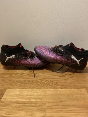 Puma Future  - Säljer ett par Puma Future Z fotbollsskor i svart och lila med coola randiga detaljer. Skorna har snörning, låg profil och en stretchig strumpkrage för skön passform. Yttersulan är genomskinlig med rosa och svarta inslag och Puma-logga på sidan.