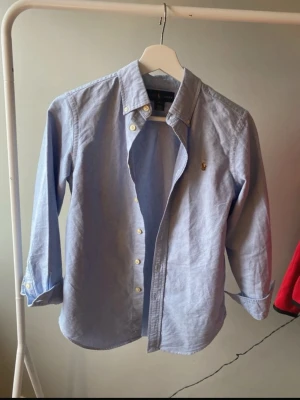 Blå skjorta från Ralph Lauren - Snygg ljusblå skjorta från Ralph Lauren med klassisk krage och gul broderad logga på bröstet. Skjortan har långa ärmar, knappar framtill och vid manschetterna. Modellen är custom fit och tillverkad i mjuk bomull som känns skön mot huden.