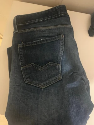 Replay blå jeans med slitningar - Säljer ett par snygga Replay jeans i blå tvätt med slitningar och detaljerade sömmar. Klassisk femficksmodell med logga på bakfickan och tydliga slitningar framtill för en cool look. Jeansen har knappgylf och är tillverkade i mjukt jeansmaterial.