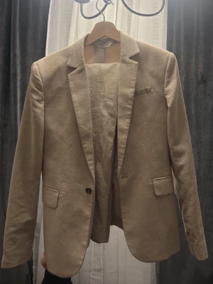Beige kostym från H&M i linne - Säljer en stilren beige kostym från H&M i linne. Kavajen har klassisk passform med två fickor med lock och en bröstficka. Byxorna är raka och matchar perfekt till kavajen. Perfekt för dig som vill ha en fräsch och luftig look.