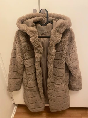 Beige pälsjacka med huva från Amisu - Säljer en fluffig beige pälsjacka från Amisu med huva och horisontella ränder. Jackan är lång och har en mysig, oversized passform som är perfekt för kalla dagar. Fodrad insida och mjukt material gör den extra skön att bära.