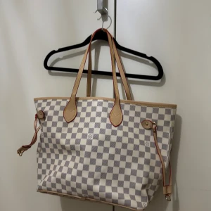 Vit väska - Säljer en vit väska med klassiskt rutigt mönster i vitt och grått. Väskan har ljusbruna skinnhandtag med röda detaljer och guldfärgade metalldelar. Perfekt storlek för att få plats med allt du behöver och har snygga läderdetaljer på sidorna.