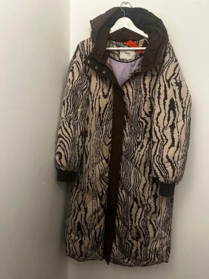 Beige och brun quiltad dunjacka från Nümph - Säljer en lång, quiltad dunjacka från Nümph med beige och brunt mönster som påminner om trästruktur. Jackan har stor huva, bruna muddar och dragkedja framtill. Materialet är polyester och jackan är perfekt för kalla dagar.