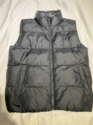 Tommy Hilfiger Essential Padded Vest i svart polyester - Säljer en svart dunväst från Tommy Hilfiger med hög krage och diskret logga på bröstet. Västen har två sidofickor och dragkedja framtill. Materialet är syntet och polyester, perfekt för kyliga dagar. Sprillans ny och ej använd!
