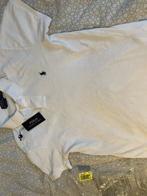 Vit pikétröja från Polo Ralph Lauren - Vit pikétröja från Polo Ralph Lauren i slim fit-modell. Klassisk krage, korta ärmar och broderad marinblå logga på bröstet. Tillverkad i bomull för en skön och luftig känsla. Perfekt för en clean och stilren look.