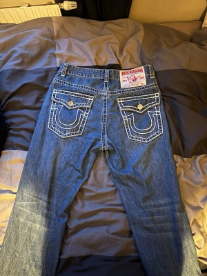 True Religion blå jeans med kontrastsömmar - Säljer ett par klassiska blå jeans från True Religion med tydliga vita kontrastsömmar och stora bakfickor med lock och knapp. Jeansen har rak passform och är tillverkade i slitstarkt denim. Märkespatch bak i midjan och detaljerade sömmar ger en unik look.