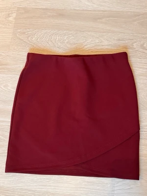 Vinröd omlottkjol i polyester - Säljer en vinröd kortkjol med omlottdetalj i nederkant. Kjolen har en enkel och stilren design, perfekt för dig som gillar att sticka ut med färg. Materialet är mjukt och stretchigt polyester som sitter skönt på kroppen. Passformen är normal och kjolen är lätt att matcha med olika toppar.