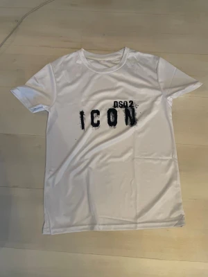 Vit ICON t-shirt med tryck - Säljer en fräsch vit t-shirt från OSO2 med coolt svart tryck där det står 'ICON' på bröstet. T-shirten har rund hals och korta ärmar, perfekt för dig som gillar enkel men snygg streetstil. Materialet känns mjukt och lätt mot huden.