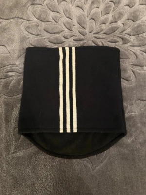 Svart Adidas halsvärmare med ränder - Säljer en svart halsvärmare från Adidas med tre vita ränder på ena sidan och Adidas-logga på andra. Materialet är mjukt och känns som fleece, perfekt för kalla dagar. Enkel och stilren design som passar till träning eller utomhusaktiviteter.