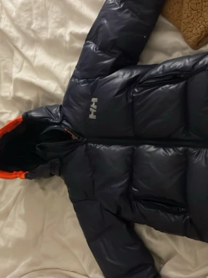 Mörkblå pufferjacka från Helly Hansen - Säljer en mörkblå pufferjacka från Helly Hansen med orange detaljer på insidan av huvan. Jackan har dragkedja framtill och två sidofickor. Perfekt för kalla dagar och har en riktigt schysst streetstil. Märket syns tydligt på bröstet och insidan.finns en liten reva vid armen 