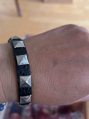 Svart läderarmband med nitar - Snyggt svart armband i skinn med silvriga pyramidformade nitar längs hela bandet. Armbandet har ett klassiskt spänne i metall som gör det enkelt att justera storleken. Perfekt accessoar för dig som gillar en edgy stil.