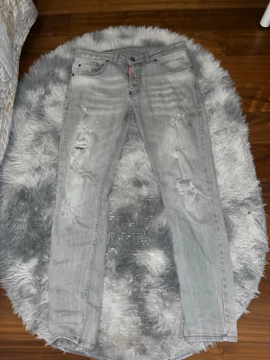 Grå Dsquared2 jeans med slitningar - Säljer ett par grå jeans från Dsquared2 i storlek 46. De har coola slitningar och detaljer på framsidan, klassisk femficksdesign och en snygg Dsquared2-label bak. Jeansen är raka i modellen och har en normal passform. Materialet är jeans med en mjuk känsla.