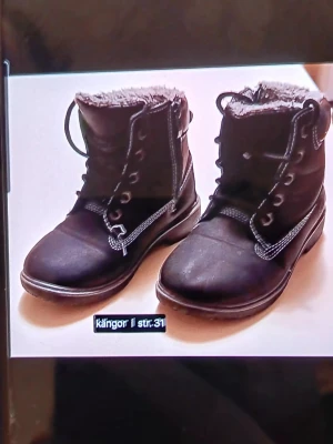 Svarta kängor med snörning strl 31 - Säljer ett par svarta kängor i storlek 31 med snörning och mjukt foder. Skorna har rund tå och grov sula, perfekta för kalla dagar. Klassisk design med kontrasterande sömmar och dragkedja på sidan för enkel på- och avtagning. Perfekt inför vintern nu.