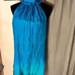Blå halterneckklänning med volang - Supersnygg blå halterneckklänning i glansigt material med volangdetalj runt halsen och knäppning bak. Klänningen har en loose passform och är ärmlös, med en cool ombre-effekt från blått till mörkare ton nedtill. Perfekt för sommarens alla tillfällen!