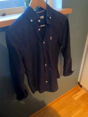 Mörkblå skjorta från U.S. Polo Assn. - Snygg mörkblå skjorta från U.S. Polo Assn. i storlek S. Skjortan har klassisk krage med knappar, broderad logga på bröstet och långa ärmar. Tillverkad i mjuk bomull som känns skön mot huden. Perfekt för dig som gillar stilrena och tidlösa plagg.