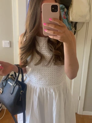 Vit ärmlös sommarklänning från Gant - Superfin vit klänning med ärmlös design och broderade hål upptill. Kjolen är lätt utställd och ger en luftig känsla, perfekt för varma dagar. Klänningen har en klassisk och fräsch look som passar till många tillfällen.