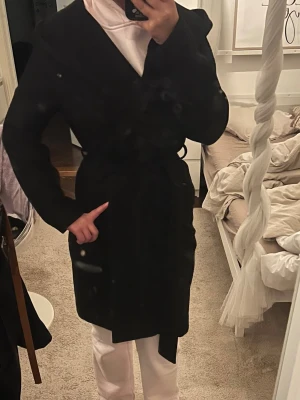 Svart kappa med bälte - Säljer en svart kappa med bälte i midjan och omlottmodell. Jackan har lång ärm och en klassisk, stilren look som passar perfekt till höst och vinter. Enkel att matcha med olika outfits och ger en snygg siluett.