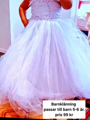 Vit långklänning med rosett - Superfin vit långklänning för barn med fluffig tyllkjol och pärldetaljer upptill. Klänningen har breda axelband och en stor rosett baktill i midjan. Perfekt för speciella tillfällen och ger en riktigt prinsesskänsla.