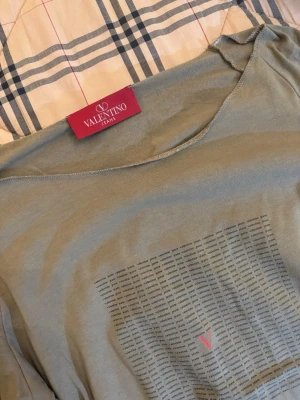 Grön topp från Valentino Jeans - Snygg grön topp från Valentino Jeans med tryckt mönster av små svarta textrader och ett rött V på framsidan. Rund halsringning och mjukt material som känns skönt mot huden. Perfekt för dig som gillar stilrena designer med en twist.