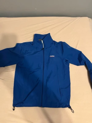Windbreaker Peak Performance jacka - Snygg blå vindjacka från Peak Performance med hög krage och hel dragkedja framtill. Jackan har justerbara snoddar i nederkant och diskret logga på bröstet. Perfekt för blåsiga dagar och enkel att matcha med jeans eller träningskläder.