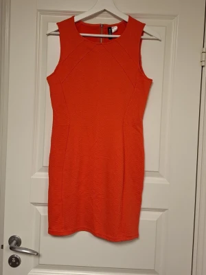 Orange kortklänning från H&M Divided - Säljer en snygg orange kortklänning från H&M Divided i storlek 38. Klänningen är ärmlös, har en figurnära passform och en diskret struktur i tyget. Dragkedja bak för enkel påklädning. Perfekt för dig som vill sticka ut!
