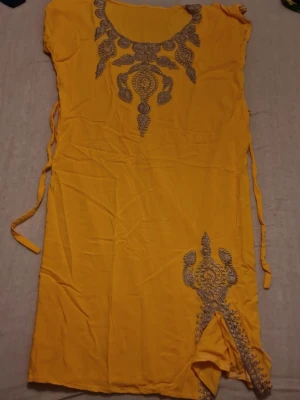Gul blus med broderi och knytband - Säljer en gul blus med detaljerat silvrigt broderi runt halsringningen och nedtill. Blusen har knytband i sidorna och en liten slits framtill. Materialet känns lätt och luftigt, perfekt för varmare dagar. Unik och färgstark topp som sticker ut. Denna klänning är köpt i Tunisien, den kallas för "kaftan". En traditionell tunisisk klänning som används eftersom det är mycket varmt i det landet. Perfekt för en resa till Tunisien om du vill respektera kulturen eller om du själv är tunisier🫶