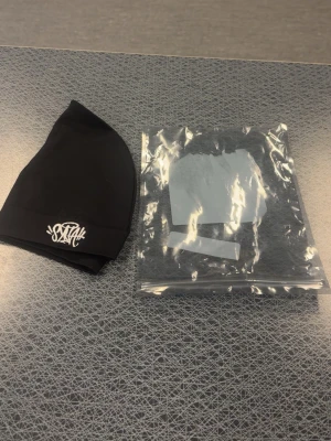 Svart mössa från Syna world  - Säljer en svart mössa från Syna world med vit broderad logframtill. Mössan är enkel och stilren i formen, tillverkad i mjuk bomull som sitter skönt på huvudet. Perfekt accessoar för kalla dagar och streetwear-stil