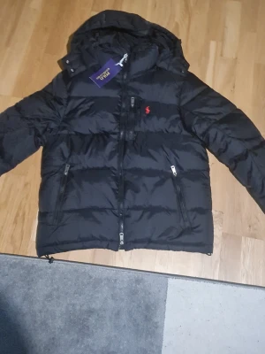 Svart pufferjacka från Polo Ralph Lauren - Svart pufferjacka från Polo Ralph Lauren med huva och flera dragkedjor. Jackan har det klassiska röda Polo-logot på bröstet och är tillverkad i polyester. Perfekt för kalla dagar och har en snygg, sportig look.