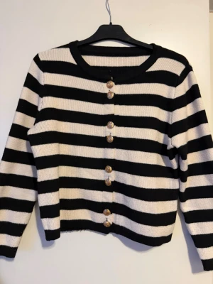 Randig svartvit kofta med guldknappar - Säljer en snygg kofta med breda svarta och vita ränder. Koftan har långa ärmar och är dekorerad med sex guldiga knappar framtill. Materialet är stickat och ger en mjuk och skön känsla. Perfekt för dig som gillar klassisk stil med en twist.