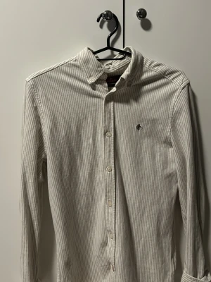 Randig skjorta från Morris XS - Snygg vit och beige randig skjorta från Morris i storlek XS. Skjortan har klassisk krage, knappar hela vägen och lång ärm. Materialet är mjukt och känns som bomull. Diskret Morris-logga broderad på bröstet och etikett i ärmslutet.