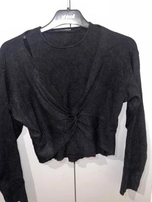 Svart croppad kofta från Kooiook - Svart ribbad kofta från Kooiook med lång ärm och croppad passform. Framsidan har en snygg twist-detalj som ger en cool look. Perfekt att styla med höga jeans eller kjol. Materialet är stickat och känns mjukt mot huden.