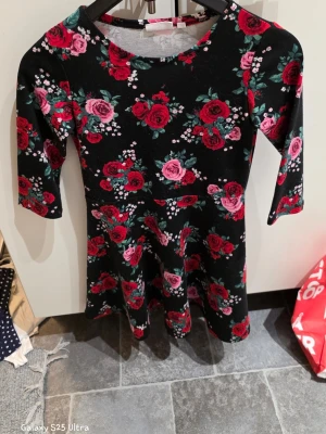 Blommig svart klänning från KappAhl - Svart kortklänning från KappAhl med färgglatt blommigt mönster i rött och rosa. Klänningen har rund hals, trekvartsärm och ett mjukt, stretchigt material som känns bekvämt. Perfekt för dig som gillar romantiska och färgstarka plagg.