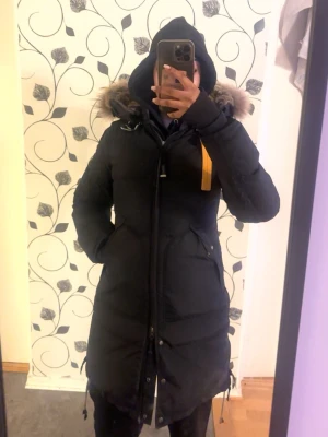 Svart lång dunjacka Parajumpers - Säljer en svart lång dunjacka från Parajumpers med huva och pälsdetalj. Jackan har dragkedja, knappar och en gul rem vid halsen. Perfekt för kalla dagar och riktigt snygg passform. Klassisk patch på ärmen och flera praktiska fickor.