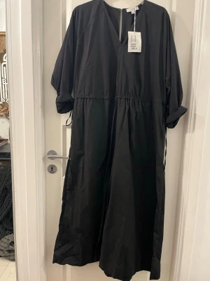 Svart långklänning från COS - Svart långklänning från COS med v-ringad hals och långa ärmar. Klänningen har en avslappnad passform med knyt i midjan och är tillverkad i mjuk bomull. Perfekt för dig som gillar minimalistisk stil och vill ha något enkelt men snyggt.