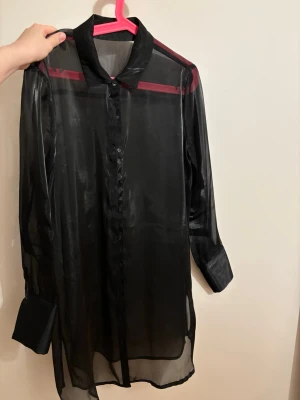Svart transparent skjorta Gina Tricot - Säljer en svart transparent skjorta från Gina Tricot i storlek 38. Skjortan har lång ärm, klassisk krage och knappar hela vägen framtill. Materialet är tunt och glansigt, vilket ger en cool och edgy vibe. Perfekt att styla lager på lager eller med ett linne under.