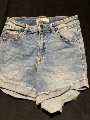 High waist jeansshorts ljusblå - Säljer ett par ljusblå high waist jeansshorts i klassisk femficksmodell. Shortsen har råa, fransiga kanter och tydliga sömmar i kontrastfärg. Perfekta för sommaren och ger en avslappnad vibe till din outfit.