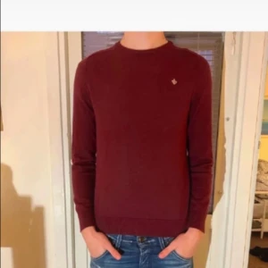 Vinröd stickad tröja med broderad logga - Vinröd stickad tröja från Morris med rund hals och diskret broderad logga på bröstet. Tröjan har lång ärm och ribbade muddar vid ärmslut och nederkant. Perfekt för dig som gillar stilren och klassisk look. Tröjan är i storlek M men skulle säga att den sitter som en S