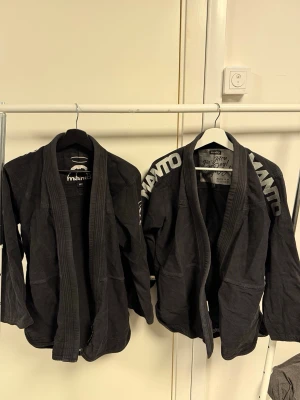 Svarta Manto Jiu Jitsu jackor - Två svarta Jiu Jitsu jackor från Manto med vit text på axlarna och ryggen. Jackorna har klassisk krage och är tillverkade i slitstarkt bomullstyg. På ryggen står det 'TRAIN JIU JITSU GO TO HEAVEN' i vitt. Perfekt för dig som tränar brasiliansk jiu jitsu.
